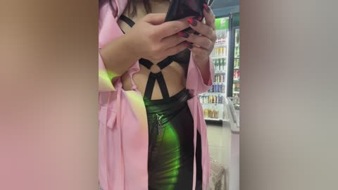 Media: A video of a woman with light skin and long dark hair, wearing a black strappy bra, green leather pants, and a pink robe, taking a selfie in a brightly lit convenience store.