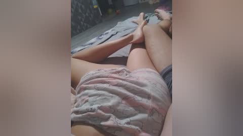 A video of a woman lying on her side in a dimly lit bedroom, wearing floral pajamas. Her legs are crossed at the ankles, and her head is turned towards the camera.