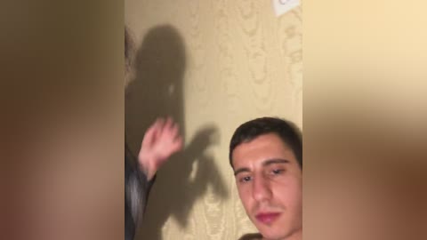 Video of a young man with short dark hair, wearing a black shirt, standing against a beige wallpaper with a floral pattern, casting a shadow on the wall.
