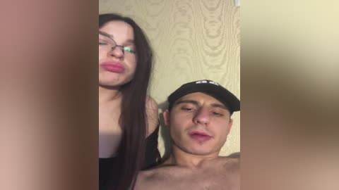 Video of a young woman with long brown hair and glasses, and a young man with a cap, both kissing. They stand in front of a textured, pale yellow wall.