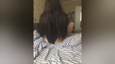 Video of a woman with long, straight, dark brown hair, sitting on a bed with striped sheets, facing away, in a dimly lit bedroom with beige walls and a white cabinet in the background.