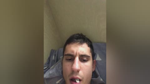 Video of a young man with short dark hair, lying on a bed, eyes closed, mouth slightly open, with a visible cigarette in his mouth. The room has beige walls and a gray bedspread.