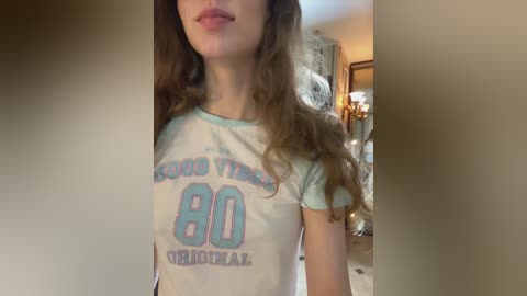 Video of a young woman with long brown hair, wearing a white T-shirt with \"Cloud 80 Original\" text, in a dimly lit, ornately decorated room with a chandelier and plush furniture.