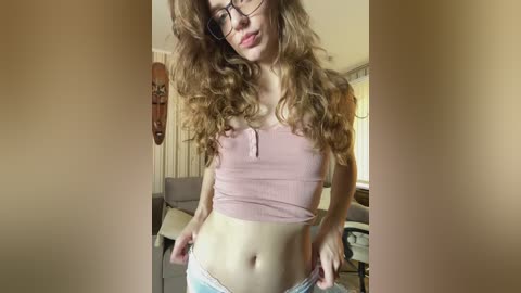 A video of a young woman with long, curly brown hair, wearing glasses, a pink crop top, and white panties, standing in a bright, modern living room with a wooden wall and window.