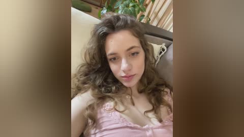 A video of a young Caucasian woman with long, wavy brown hair, wearing a pink lace camisole, sitting on a beige couch, with a green potted plant in the background.
