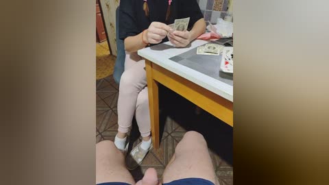 Video of a woman in a black top, light pink pants, and white shoes sitting at a table with a stack of dollar bills and playing cards, viewed from the feet of a man in blue shorts.