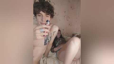 Media: Video of a shirtless, curly-haired man with a tattoo on his arm, holding a lighter, sitting on a bed with a woman in a black dress behind him, in a dimly lit, beige room.