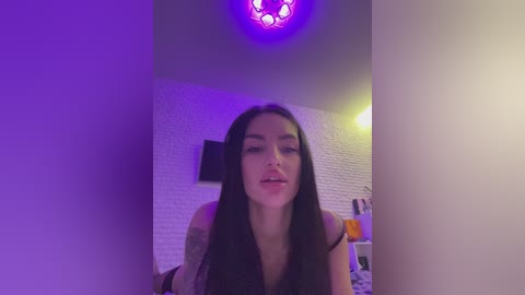 Media: Video of a young woman with long black hair, fair skin, and a slender physique, wearing a sleeveless top, standing in a dimly lit room with purple LED lighting and white brick walls.