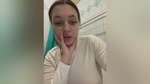 Media: A candid video of a young woman with light skin and dark hair, wearing a beige long-sleeved top, resting her head on her hand, in a tiled bathroom with green and white accents.