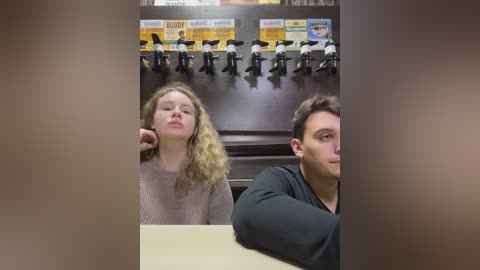 Media: Video of two people in an office setting. A blonde woman with curly hair and a man with short dark hair sit side by side, both looking at a display of toy figures.