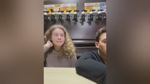 Media: A video captures a young woman with curly blonde hair and a casual beige sweater, looking contemplative, sitting in a diner with a man in a black shirt. The background features a row of black and white checkered booths and a colorful sign.