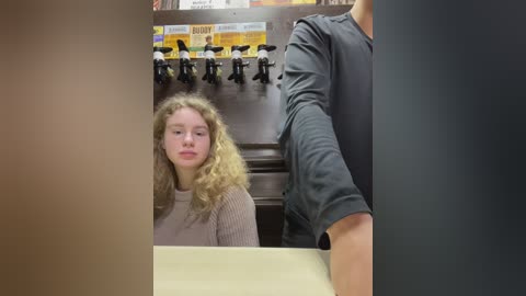 Media: Video of a blonde woman with curly hair wearing a beige sweater, seated at a coffee counter, facing a man in a black shirt pouring coffee from a dispenser with various coffee options displayed above.