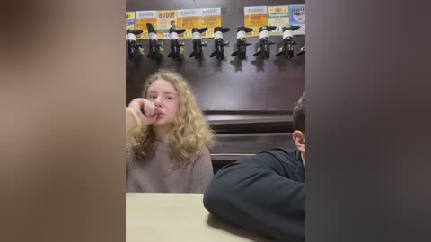 Media: Video of a blonde girl in a beige sweater eating a banana in a school cafeteria with black-and-white name tags on the wall.