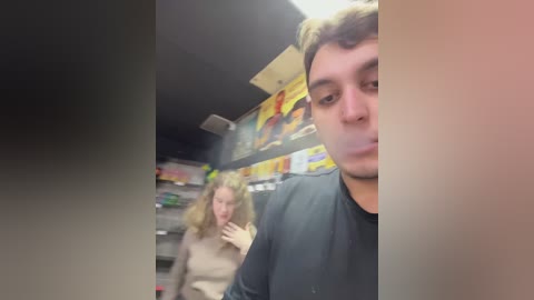 Media: Video of a blurry, slightly out-of-focus woman with shoulder-length, wavy blonde hair, wearing a beige sweater, taking a selfie in a dimly lit convenience store. The background features colorful, abstract wall art and shelves with various products.