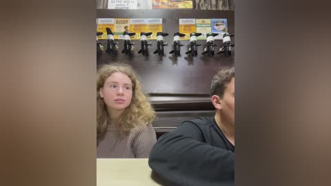 Media: Video of a woman with curly blonde hair in a gray sweater and a man with short brown hair in a black shirt, seated at a table, surrounded by a wall-mounted beer tap display.