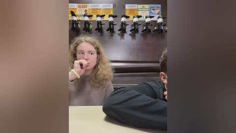Media: Video of a woman with curly hair, wearing a light sweater, sitting at a table. Behind her, a row of uniformed dolls is displayed in a fast-food restaurant.