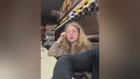 Media: Video of a young woman with curly blonde hair, wearing a beige sweater and dark pants, sitting in a bowling alley with a colorful, checkered ceiling and arcade games in the background.