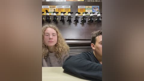 Media: Video of a young woman with curly hair, eyes closed, sitting next to a man with short hair, both in casual attire. Background shows a restaurant with toy figurines on shelves.
