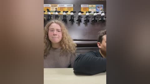 Media: Video of a woman with curly hair, wearing a grey sweater, seated at a table in a dimly lit bar, surrounded by black bar stools and beer posters.