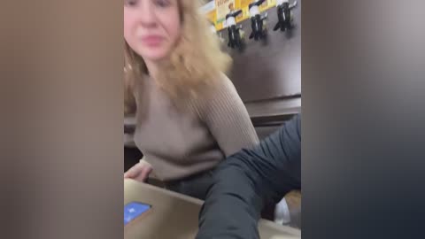 Media: Video of a blonde woman in a beige sweater and dark pants, sitting on a beige box in a dimly lit room with hanging yellow bags and a shelf.