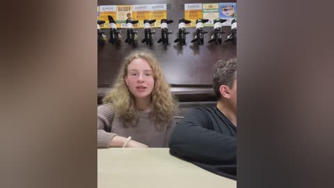 Media: Video of a young blonde woman with curly hair in a beige sweater, seated at a table in a restaurant, surrounded by people watching a TV screen displaying sports highlights.
