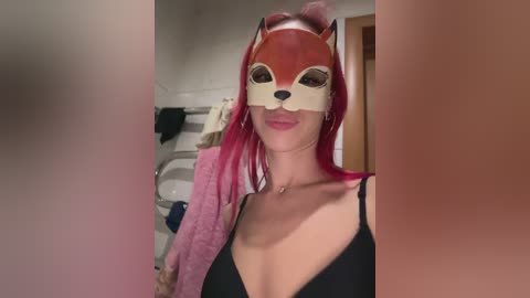 Video of a woman with long pink hair wearing a red fox mask and a black lace bra, standing in a bathroom with white tiles and a towel rack.