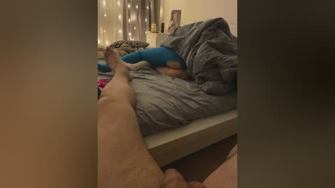 Media: A video of a messy bedroom with a bed covered in blankets and pillows, featuring a person's bare legs and feet, and a string of fairy lights in the background.