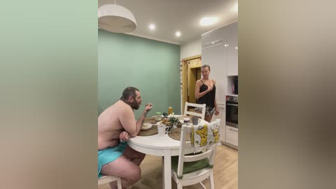 Media: Video of a chubby, shirtless man in blue shorts, sitting at a table, eating a sandwich, while a woman in a black dress watches him from the background, in a modern, light-colored kitchen.