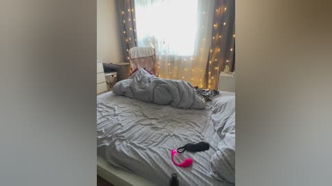 Media: Video of a messy bedroom with a grey bedspread, a pink vibrator on the bed, and sheer curtains with fairy lights.