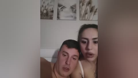 Video of a man with short dark hair and a woman with long dark hair, both sitting on a white couch, looking at each other intimately.