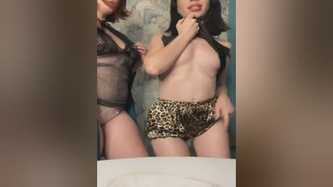 Media: A video of two women, one in a sheer black bralette and leopard-print shorts, the other in a black bralette, standing in a bathroom.