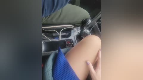 Video of a woman in a blue dress sitting in a car, with her hand on her thigh, showing a glimpse of her pale skin. The car interior is modern, with a dashboard and steering wheel visible.