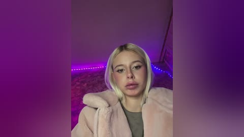 Video of a young, pale-skinned woman with platinum blonde hair, wearing a pink fuzzy coat, standing in a dimly lit room with purple LED strip lighting.