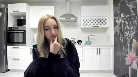 Media: Video of a blonde woman in a black robe, sitting in a modern white kitchen with stainless steel appliances and a chalkboard wall.