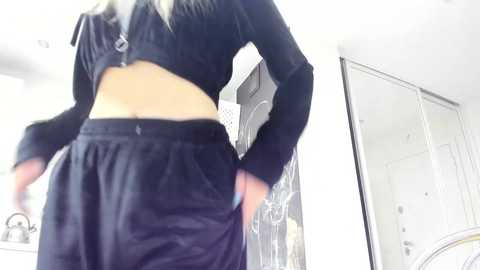 Media: Video of a slender woman with long, straight blonde hair, wearing a black crop top and high-waisted black pants, standing in a bright, modern bedroom with white walls and a large mirror.