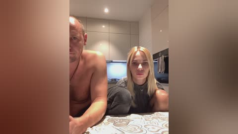 Media: Video of a muscular, shirtless man with a bald head and a blonde woman with long hair, both smiling, in a modern kitchen with sleek cabinets and stainless steel appliances.