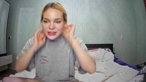 Media: A video of a blonde woman with fair skin and a grey shirt, animatedly touching her ears in a bedroom with light blue, textured wallpaper.