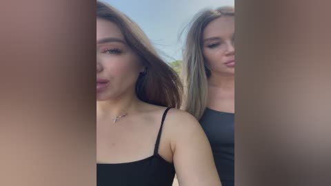 Media: Video of two young women with fair skin and long brown hair, wearing black spaghetti-strap tops, standing closely together outdoors.