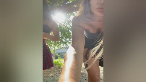 Media: Video of two women outdoors, one with long blonde hair, wearing a black top, bending forward; the other, partially visible, in a plaid skirt. Sunlight filters through leaves, creating a bright, summery atmosphere.