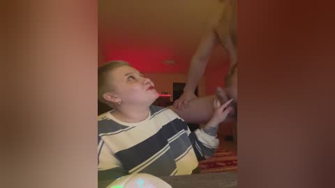 A video depicts a blonde woman in a striped shirt, kneeling, with a bald man above her, in a dimly lit room with a red light and a patterned rug.