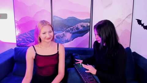 Video of two women in a modern living room. One blonde, wearing a sheer red top, sits on a blue couch; another brunette with long hair, in black, uses a keyboard. Background features digital art of a mountainous landscape.