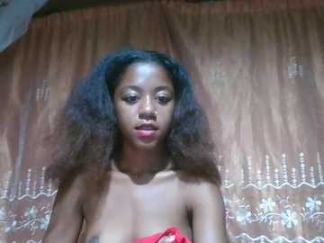 Video of a young Black woman with voluminous, curly hair and full lips, wearing a strapless red dress. She stands against a backdrop of beige curtains with ornate silver designs.