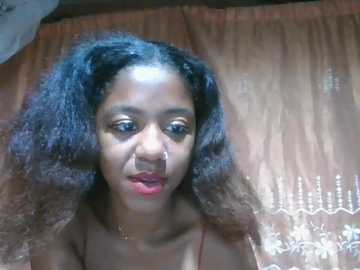 Video of a young Black woman with medium skin tone and long, curly black hair. She wears a red spaghetti-strap top, has a nose stud, and sits on a patterned, beige bedspread.