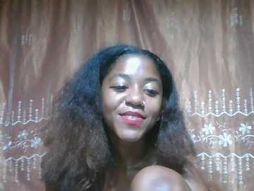 Video of a smiling, light-skinned Black woman with curly hair, wearing a patterned, brown and white dress. Background shows a beige curtain with decorative silver embroidery.