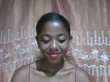 A video of a smiling young woman with medium brown skin, dark hair pulled back, wearing a red spaghetti strap top against a peach satin backdrop adorned with white floral embroidery.
