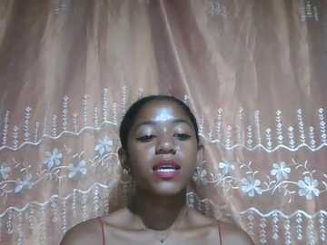 Video of a young Black woman with medium brown skin and dark hair, wearing a red top, against a backdrop of a sheer, light pink curtain adorned with white floral embroidery.