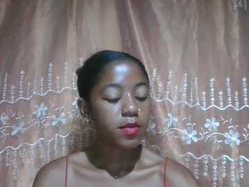 Video of a serene African-American woman with medium brown skin and straightened dark hair, wearing a red spaghetti-strap top, standing against a backdrop of shimmering, gold, floral-patterned fabric.