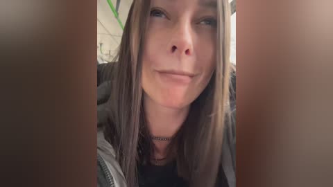 Media: Video of a smiling young woman with long, straight brown hair, wearing a black top and gray jacket, taken indoors with blurry background.