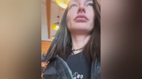 Media: Video of a young Caucasian woman with straight brown hair, light skin, and blue eyes, wearing a black T-shirt and a dark jacket. The background shows a modern, wooden-beamed room with warm lighting.
