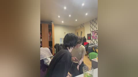 A video of a casual indoor gathering with several people, including one in a black jacket, sitting around a table. The background features a beige wall, a bookshelf, and a cluttered room.